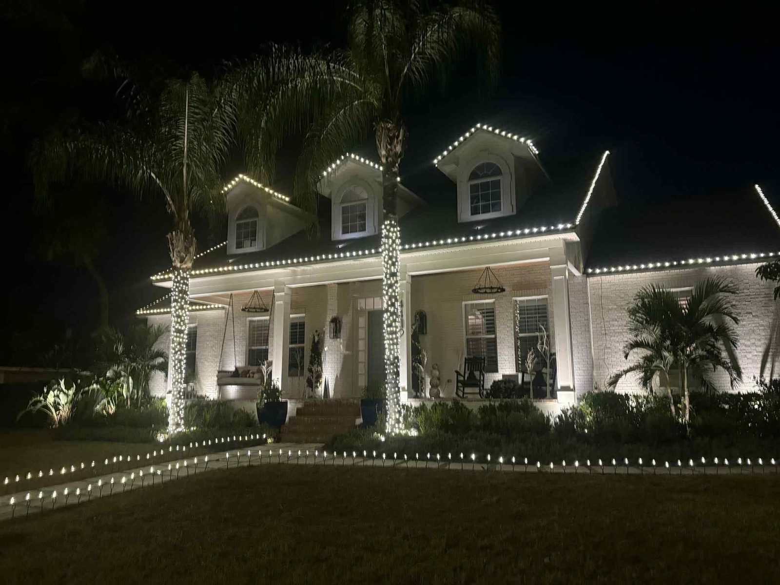 A finished install on a white brick house. Bulbs trace the roofline and all three dormers, the porch columns and palms are wrapped, and pathway lights run the length of the walk.