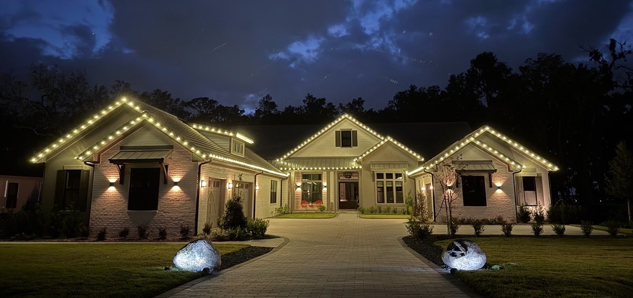 A house we lit, photographed at dusk. Warm bulbs run the full roofline and every gable, with the driveway and entry lit below.